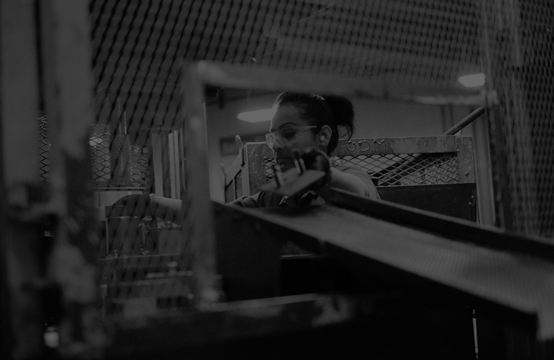 A person wearing safety glasses operates industrial machinery with a conveyor system and metal fencing in a factory, working on large bed stamping of automotive stamped parts.