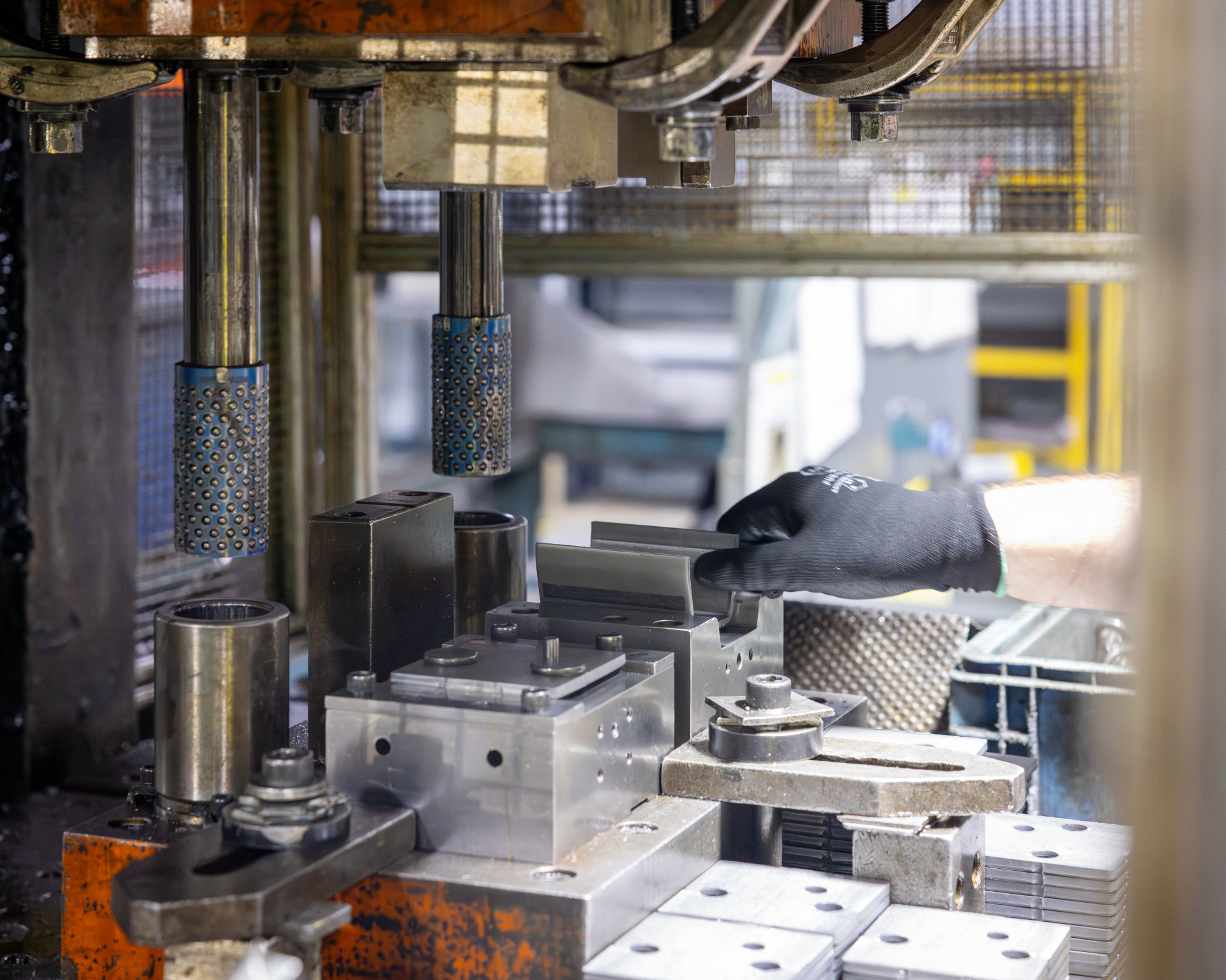 A gloved hand adjusts metal components inside a heavy gauge stamping machine press in a factory setting, surrounded by various mechanical parts and equipment.